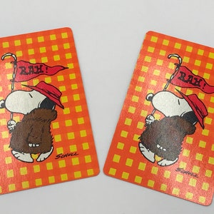 May include: Two playing cards featuring Snoopy in a brown outfit and a red hat, holding a flag that says "RAH!". The cards have an orange and yellow checkered background. The artist's signature, "Schulz", is visible.