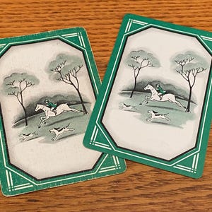May include: Two playing cards with a green border and a hunting scene. Each card features a rider on a white horse, two dogs, and bare trees. The background is a light green color.
