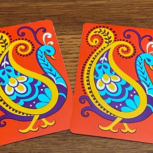 May include: Two playing cards with a vibrant peacock design. The cards feature a red background with a yellow, purple, and turquoise peacock illustration. The peacock has a paisley-like body and a curved neck.