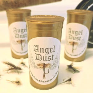 May include: Three gold-colored cylindrical containers labeled "Angel Dust" with a dragonfly graphic. Several artificial flies are scattered in front of the containers. The containers are on a white surface.