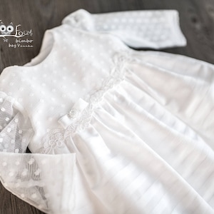 May include: A white baby dress with a delicate design. The dress features a dotted pattern on the sleeves and bodice, with a bow and lace detailing. The dress is made of a soft, lightweight fabric, perfect for a special occasion.