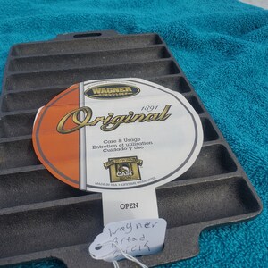 May include: A black cast iron bread pan with 12 individual slots. The pan has a white label with the text "Wagner Original 1891" and "Care & Usage, Entretien et utilisation, Cuidado y Uso". The label also has a logo that says "Award Winning Cast Iron" and "Made in USA - Lifetime Guarantee".