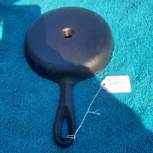 May include: A black cast iron skillet lid with a handle and a hole in the center. The lid is round and has a slightly textured surface. A white tag with the text "BSR 65/11e" is attached to the handle with a string. The lid is on a blue surface.