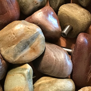 May include: Close-up of a collection of polished river stones in various colors, including red, brown, gray, and tan. Some stones have metal hardware attached, suggesting they are decorative cabinet knobs or pulls.