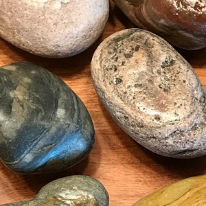 May include: Close-up of several smooth, rounded stones in various colors, including gray, brown, and tan. The stones have natural textures and patterns, and are arranged on a wooden surface. The stones vary in size and shape.