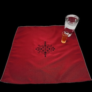 May include: A red cloth table runner with a black embroidered design. The design features a cross with a star at each point. Two glasses are on the table runner, one with water and one with a dark liquid.