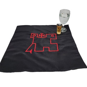 May include: A black cloth with a red embroidered design of a castle with a cross on top. There are two glasses on the cloth, one filled with water and the other with a golden liquid.
