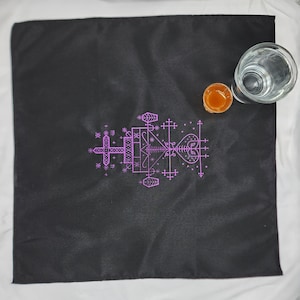 May include: A black cloth with a pink sigil design. The sigil is a complex geometric pattern with crosses, hearts, and other symbols. The cloth is laid out flat on a white surface.