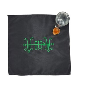 May include: A black cloth with a green sigil embroidered on it. A glass of water and a small glass of liquid are on the cloth.
