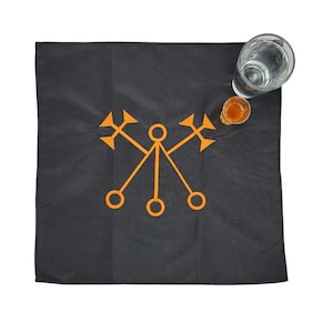 May include: A black cloth with an orange symbol of three circles connected by lines with arrows at the ends. Two glasses are on the cloth, one is clear and the other is amber.