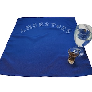 May include: A blue cloth table runner with the word "ANCESTORS" embroidered in white thread. A glass of water and a shot glass are on the table runner.
