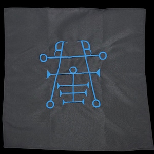 May include: A square, dark gray cloth with a blue, geometric sigil design embroidered in the center. The sigil features lines, circles, and arrow-like shapes, creating a symmetrical pattern. The cloth is set against a black background.