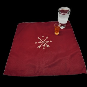 May include: A burgundy square cloth with an embroidered design of arrows and stars, with a shot glass of amber liquid and a glass of milky liquid. The cloth is likely a decorative element for serving drinks.