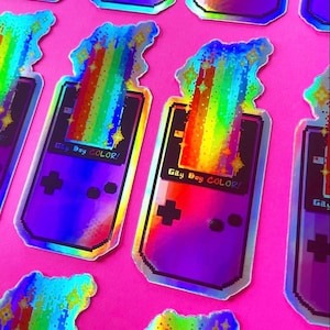 May include: A holographic sticker of a purple Game Boy Color with a rainbow pixelated design and the text "Gay Boy COLOR!"