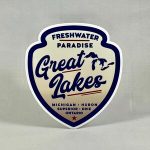 Może przedstawiać: Biała naklejka z niebieską obwódką w kształcie tarczy. Naklejka zawiera napis "Freshwater Paradise Great Lakes" w niebieskim, ozdobnym stylu pisma z czerwonym podkreśleniem. Naklejka zawiera również zarys Wielkich Jezior i napis "Michigan, Huron, Superior, Erie, Ontario" mniejszą czcionką.