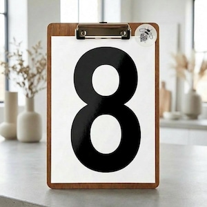 Op de afbeelding: Houten clipboard met een wit papier met een groot zwart cijfer 8. Een kleine sticker met een bijenillustratie is bevestigd. Het clipboard staat op een grijze aanrecht, geschikt voor interieur of organisatie.