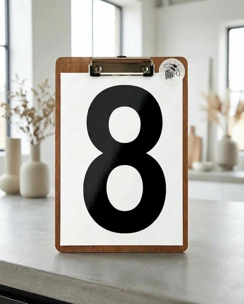 Op de afbeelding: Houten clipboard met een wit papier met een groot zwart cijfer 8. Een kleine sticker met een bijenillustratie is bevestigd. Het clipboard staat op een grijze aanrecht, geschikt voor interieur of organisatie.