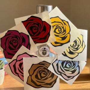 Puede incluir: Una colecci&oacute;n de pegatinas de rosas en varios colores, incluyendo rojo, amarillo, rosa y marr&oacute;n. Cada pegatina presenta un dise&ntilde;o detallado de rosa con contornos negros. Las pegatinas se muestran sobre una superficie transparente.
