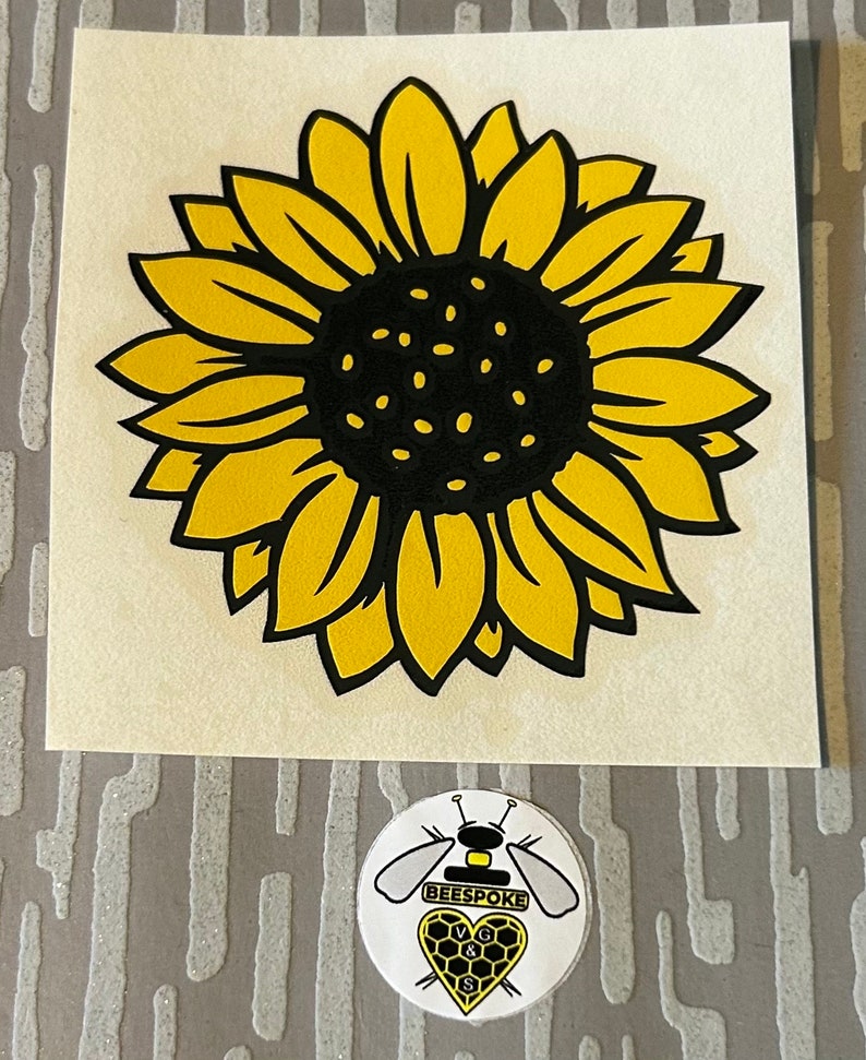May include: A yellow and black sunflower sticker with a detailed design. The sunflower has a black centre with small dots and yellow petals. Below the sunflower is a round sticker with a bee and the word "BEESPOKE".
