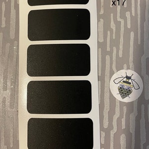 Pode incluir: Uma folha de 17 etiquetas retangulares pretas, cada uma medindo 5,08 cm x 2,54 cm, com cantos arredondados. Um pequeno adesivo redondo com uma ilustração de abelha também é visível. As etiquetas estão em um suporte branco.