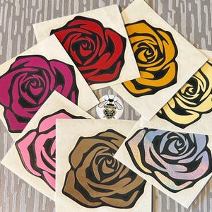 Puede incluir: Una colecci&oacute;n de pegatinas con forma de rosa en varios colores, incluyendo rojo, rosa, amarillo y marr&oacute;n, cada una con un contorno negro. Las pegatinas est&aacute;n dispuestas sobre un fondo claro, con una peque&ntilde;a pegatina circular con un logotipo de abeja.