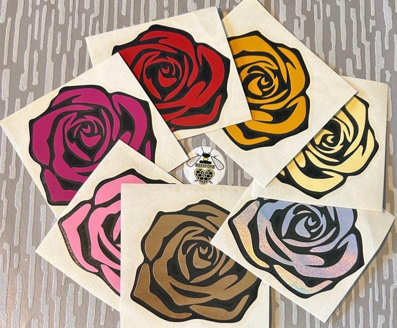 Puede incluir: Una colecci&oacute;n de pegatinas con forma de rosa en varios colores, incluyendo rojo, rosa, amarillo y marr&oacute;n, cada una con un contorno negro. Las pegatinas est&aacute;n dispuestas sobre un fondo claro, con una peque&ntilde;a pegatina circular con un logotipo de abeja.