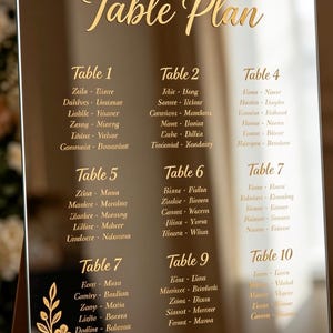 May include: A mirrored acrylic table plan with gold lettering and a floral design. The text reads "Table Plan" at the top, followed by table numbers and guest names. The date "2024.09.15" is displayed at the bottom.