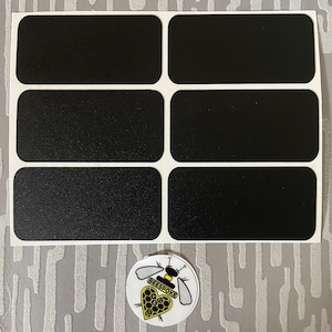 Pode incluir: Uma folha de seis etiquetas retangulares pretas com cantos arredondados em um suporte branco. Um adesivo circular com um desenho de abelha e a palavra "BEESPOKE" na parte inferior. O adesivo tem um coração amarelo com um padrão de favo de mel.