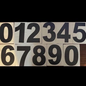 Op de afbeelding: Een set zwarte cijfers van 0 tot en met 9, gedrukt op afzonderlijke beige vierkanten. De cijfers zijn in een vetgedrukte, schreefloze lettertype, gerangschikt in twee rijen.