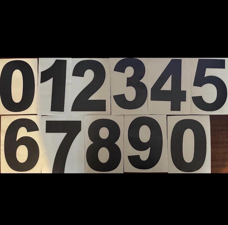 Op de afbeelding: Een set zwarte cijfers van 0 tot en met 9, gedrukt op afzonderlijke beige vierkanten. De cijfers zijn in een vetgedrukte, schreefloze lettertype, gerangschikt in twee rijen.