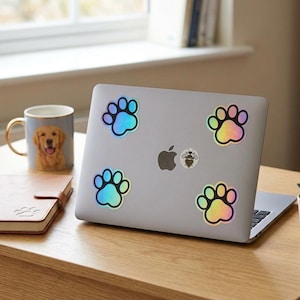 Op de afbeelding: Een zilveren laptop versierd met holografische pootafdrukstickers. Een mok met een golden retriever en een klein notitieboekje staan op een houten bureau. De stickers hebben een regenboogglans.