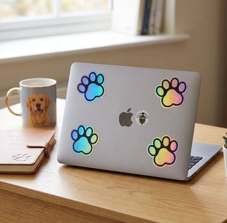 Op de afbeelding: Een zilveren laptop versierd met holografische pootafdrukstickers. Een mok met een golden retriever en een klein notitieboekje staan op een houten bureau. De stickers hebben een regenboogglans.