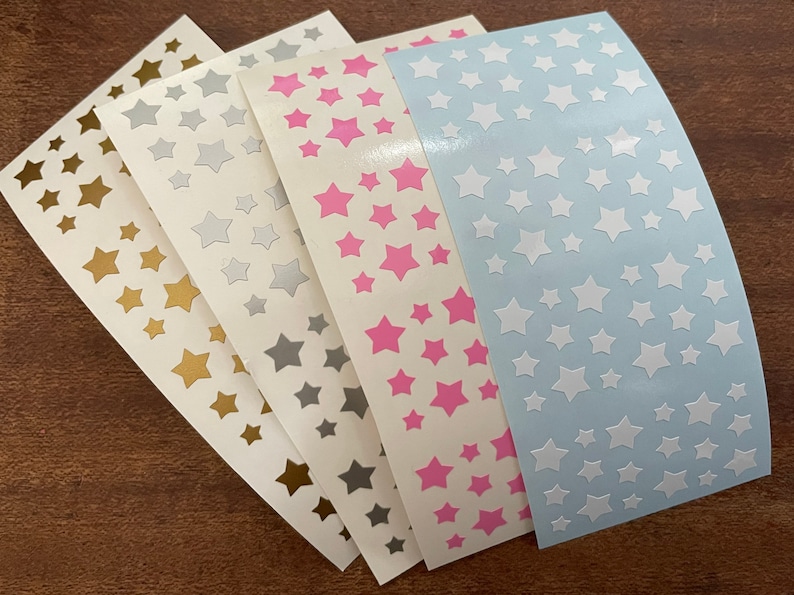 May include: A collection of star-shaped stickers in various colors, including gold, silver, pink, and white, arranged on a white background. The stickers are of different sizes and are arranged in a random pattern. The stickers are on a light blue background.