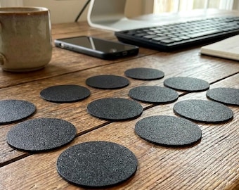 May include: A collection of black, oval-shaped coasters arranged on a wooden table. A beige mug, a smartphone, a keyboard, and a notepad are also visible. The coasters have a textured surface, suggesting they are made of a durable material.