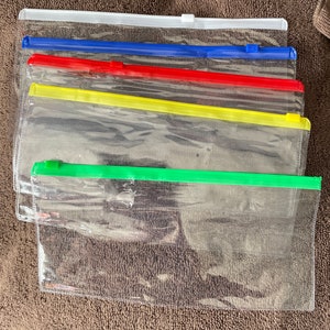 May include: A stack of clear plastic zipper pouches with colored zippers. The zippers are white, blue, red, yellow, and green. The pouches are transparent, allowing the contents to be visible.