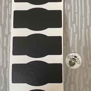 Pode incluir: Uma folha de seis etiquetas pretas de quadro-negro com bordas arredondadas. Um pequeno botão redondo com um gráfico de abelha e a palavra "Bespoke" está ao lado das etiquetas. As etiquetas estão em um fundo branco.