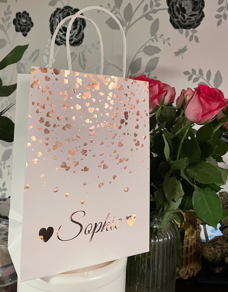 May include: White gift bag with rose gold heart confetti design and white rope handles. The name "Sophie" is printed in rose gold script with heart accents. A bouquet of pink roses is in the background.