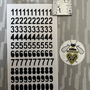 May include: A sheet of black number stickers ranging from 0 to 9, with each number repeated multiple times. A small round sticker with a bee and honeycomb design is also present. A ruler is placed next to the stickers.