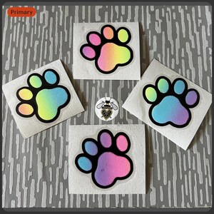 Op de afbeelding: Vier vierkante stickers met een pootafdrukontwerp. Elke pootafdruk is zwart omlijnd en gevuld met een regenboogverloop. Er is ook een kleine speld met een bijen-thema zichtbaar.