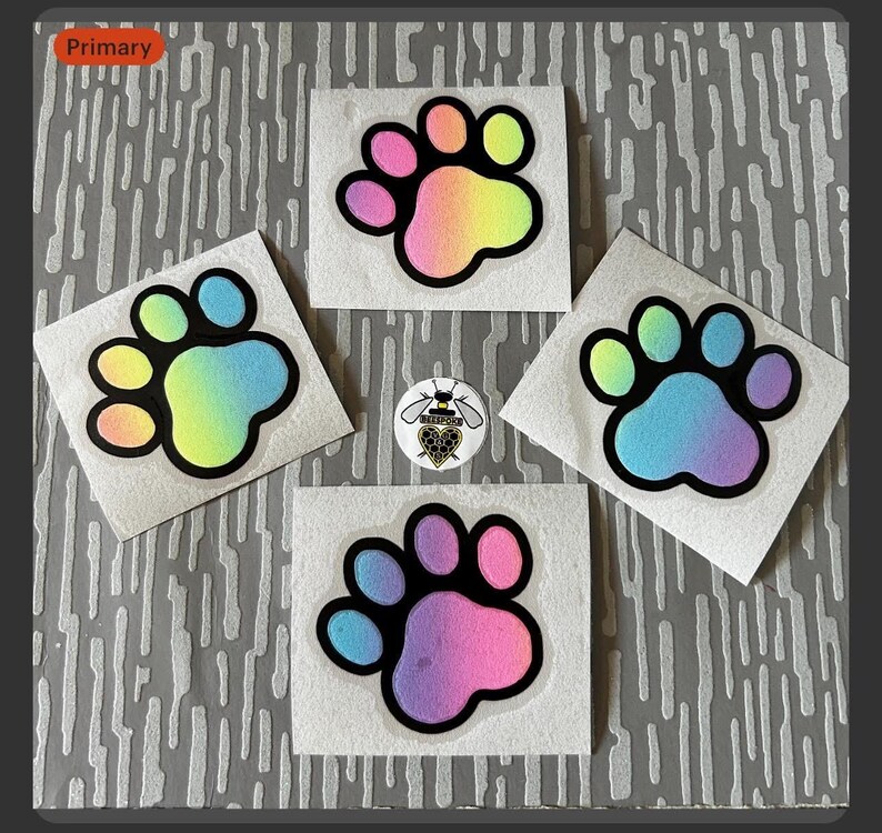 Op de afbeelding: Vier vierkante stickers met een pootafdrukontwerp. Elke pootafdruk is zwart omlijnd en gevuld met een regenboogverloop. Er is ook een kleine speld met een bijen-thema zichtbaar.
