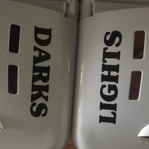 Op de afbeelding: Twee witte wasmanden met zwarte tekst. Eén leest "DARKS" en de andere "LIGHTS". Elke mand heeft rechthoekige uitsparingen en een gebogen bovenkant.