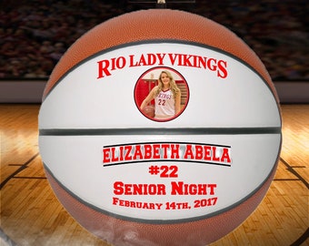 May include: A white basketball with black stripes and red text. The text reads "Rio Lady Vikings", "Elizabeth Abella #22", "Senior Night", and "February 14th, 2017". A photo of a girl in a basketball uniform is in the center of the ball.