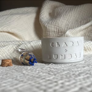 Może przedstawiać: Biały betonowy zapalacz do zapałek z wytłoczonym tekstem "Clara + Lundberg" na boku. Obok zapalacza znajduje się szklany słoik wypełniony drewnianymi zapałkami z niebieskimi końcówkami.