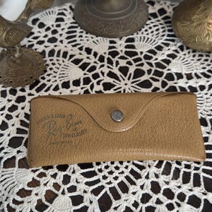 May include: A tan leather Ray-Ban sunglasses case with a snap closure. The case has the Ray-Ban logo and text printed on the front. The case is sitting on a white lace doily.