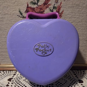 May include: A vintage Polly Pocket compact in a heart shape, lavender in color, with a matching handle. The compact features a silver oval emblem with the words "Polly Pocket" and a heart symbol. A nostalgic toy.