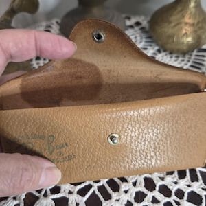 May include: Tan leather eyeglass case, open to reveal the interior. The case has a snap closure and a curved top. The brand name "Ray-Ban" is visible on the front. The case is held open by a hand.