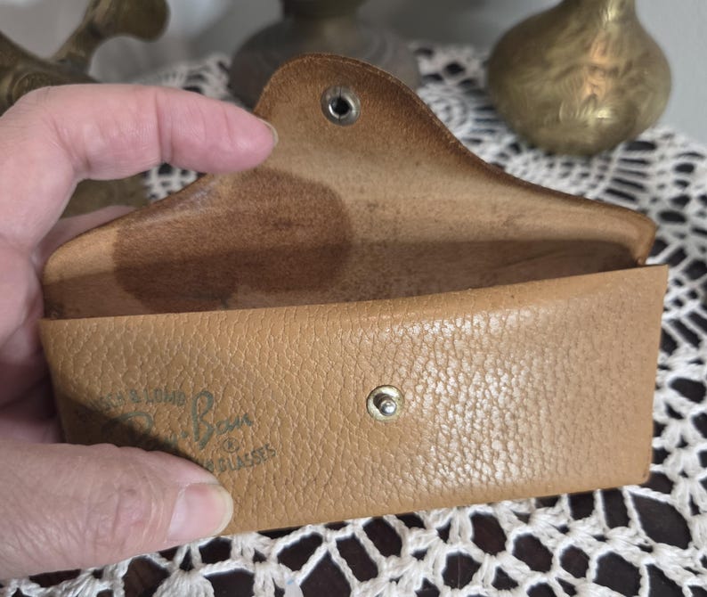 May include: Tan leather eyeglass case, open to reveal the interior. The case has a snap closure and a curved top. The brand name "Ray-Ban" is visible on the front. The case is held open by a hand.