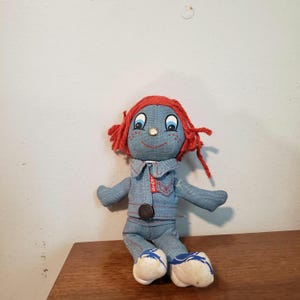 May include: A vintage denim doll with red yarn hair, blue eyes, and a smiling face. The doll wears a denim jacket and pants with white and blue shoes. A small red Levi's tag is visible on the jacket.