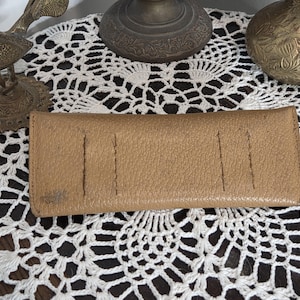 May include: A tan leather wallet with a textured surface. The wallet has several vertical stitched lines and a small stain on the front. The wallet is rectangular and appears to be closed.