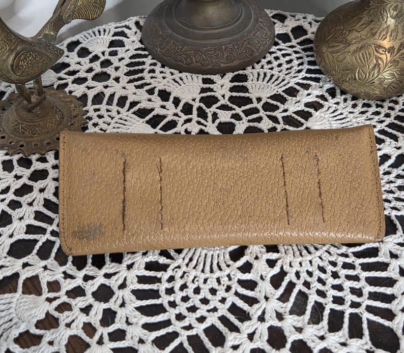 May include: A tan leather wallet with a textured surface. The wallet has several vertical stitched lines and a small stain on the front. The wallet is rectangular and appears to be closed.